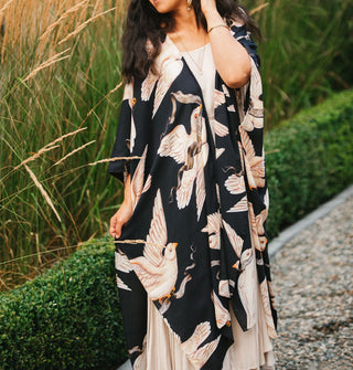 Model standing in front of a hedgerow wears a loose-fitting, black kimono-style robe with a print of large white doves carrying banners in their beaks
