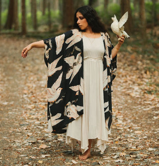 Model standing in the woods on a path covered with dead leaves wears a black robe with white doves printed on it and extends one arm while perched on her other hand is a white bird flapping its wings