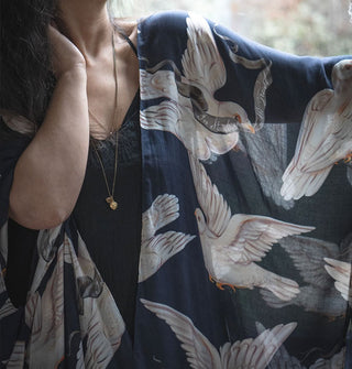 Model extends one arm to show the slightly sheer nature of the fabric of a dark blue-black robe with large white dove print that she is wearing