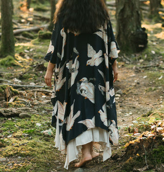 Model standing in the woods and facing away from the camera wears a flowing black kimono-style robe with print of large white doves carrying banners in their beaks