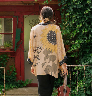 Model holding an ukulele and facing away from the camera in front of a cottage wears a cardigan with sun flower motif and contrasting black and white print borders