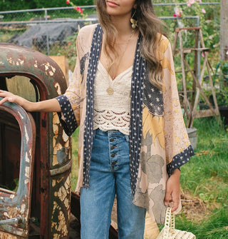 Model wears a kimono-style cardigan with sunflower motif and contrasting black and white star print borders over a crocheted top and blue button fly jeans