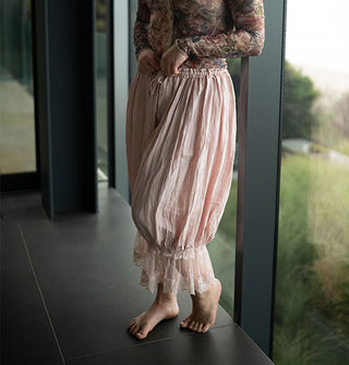 Model wears light pink pantaloons with lace trim near a floor-length window overlooking a grassy, wooded expanse