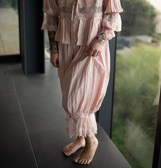 Model wears light pink pantaloons and blouse with lace trim near a floor-length window overlooking a grassy, wooded expanse