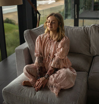 Smiling model with eyes closed sits cross-legged on a sectional sofa wearing a pink lacy blouse and pantaloon set