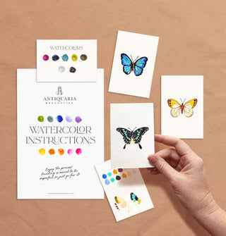 Model's hand holds a completed mini watercolor painting of a butterfly near two others and Antiquaria Mercantile watercolor kit swatch cards and instructions