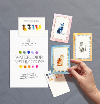 Model's hand holds one completed cat painting from an Antiquaria Mercantile watercolor kit while two others, plus a swatch card and instruction sheet rest nearby