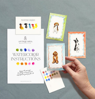 Model's hand holds one completed dog painting from an Antiquaria Mercantile watercolor kit while two others, plus a swatch card and instruction sheet rest nearby