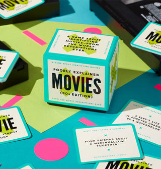 Game box of Poorly Explained Movies (80s Edition) and several pulled cards are staged on a brightly colored surface of cutout paper shapes