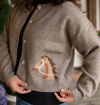 Model pulls gently at the bottom ribbed hem of a gray-brown button-down cardigan with brown horse head detail