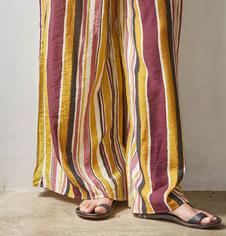 Model wears wide-legged brown, burgundy, rose, gold, and white striped pants with sandals