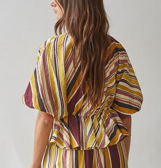 Model wears a brown, burgundy, rose, gold, and white striped top with gathered waist and puff elbow-length sleeves paired with matching pants