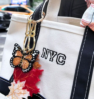 A monarch butterfly purse charm is attached to the handle of a navy and white canvas NYC tote along with maple leaves and a bear