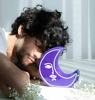Model with eyes closed leans toward a blueish-purple crescent moon-shaped vase flanked by small white flowers