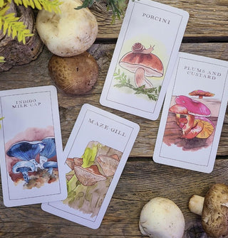 Four sample cards from the Mushroom Spotters Deck (Indigo Milk Cap, Porcini, Maze Gill, and Plums and Custard) rest on a rustic wooden surface with ferns and mushrooms