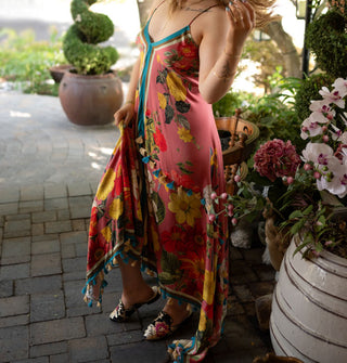 Model wears a pink dress with large floral print, asymmetrical high-low cut, contrast stripes along the neckline, bottom, and front center, and two rows of tassels, one along the bottom hem and the other at the hip