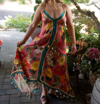 Model wears a pink dress with large floral print, asymmetrical high-low cut, contrast stripes along the neckline, bottom, and front center, and two rows of tassels, one along the bottom hem and the other at the hip