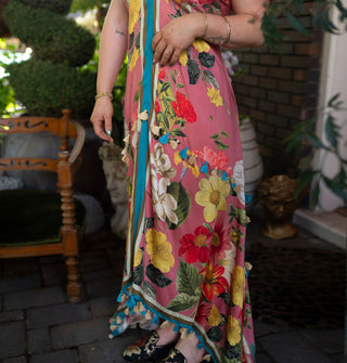 Model wears a pink dress with large floral print, asymmetrical high-low cut, contrast stripes along the bottom and down the front center, and two rows of tassels, one along the bottom hem and the other at the hip