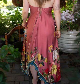 Model wears a pink dress with a tied keyhole back and enlarged, colorful floral designs along the bottom which is finished with contrast stripes and tassels along the hem