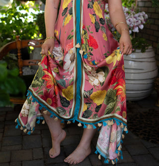 Model holds the skirt of a pink dress that features bold, colorful floral designs contrasted by center vertical and bottom hem stripes and tassels