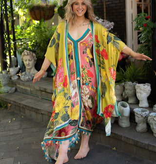Model wears a long olive-chartreuse kimono-style robe with large floral print and contrast edge stripes paired with a dress with tassels along the bottom