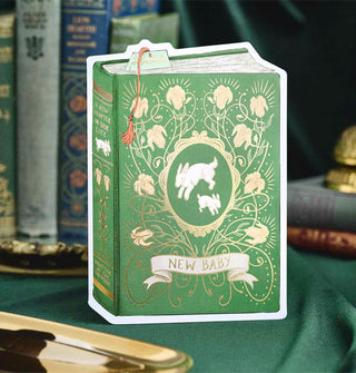 Green greeting card is designed to resemble a book with jumping white rabbits on its over inside an ornate gold floral frame above the words, "New Baby" in a banner; on its spine is the message, "A new chapter in your life written by the universe"