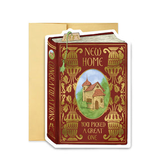 Burgundy greeting card in front of a gold envelope is designed to resemble a book cover with gold foil ornamentation surrounding a central framed illustration of a charming abode and the words, "New home" at the top and, "You picked a great one" at the bottom; on its spine are the words, "Congratulations" and, "Cozy House Publishing"