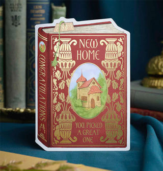 Burgundy greeting card is designed to resemble a book cover with gold foil ornamentation surrounding a central framed illustration of a charming abode and the words, "New home" at the top and, "You picked a great one" at the bottom; on its spine are the words, "Congratulations" and, "Cozy House Publishing"