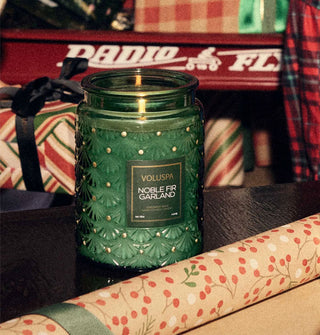 Lit Noble Fir Garland Voluspa candle in green embossed glass vessel is staged with a roll of holiday wrapping paper, a Radio Flyer wagon behind, and wrapped presents