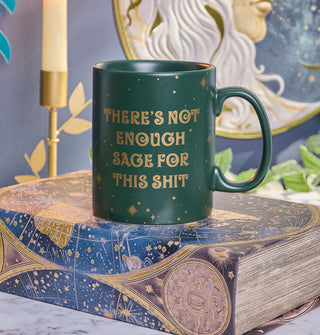 Green There's Not Enough Sage for This Shit coffee mug rests on a book with celestial map cover in front of other mystical items