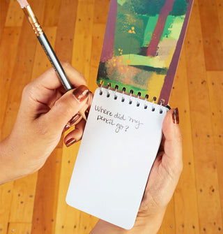 Model's hands hold a pencil and small notebook with abstract interior cover design and the words, "Where did my pencil go?" jotted on the first white page