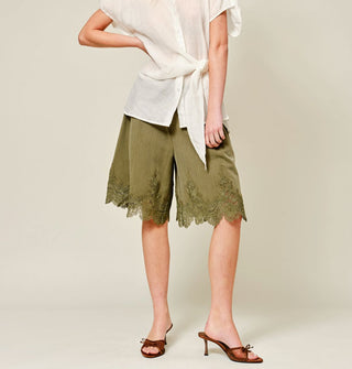 Model with hand on hip wears a loose-fitting white blouse with tie detail, olive green wide-legged shorts with lace-trimmed hems, and brown open-toed kitten heels with bow accents