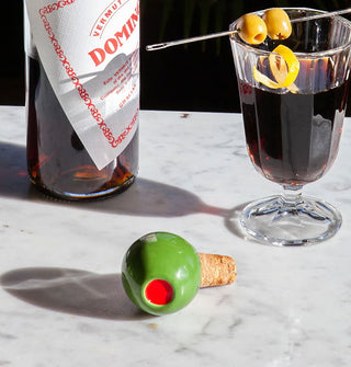 A ceramic and cork green olive bottle stoper rests on its side on a marble tabletop with liquor bottle and cocktail with olive and lemon twist garnishes