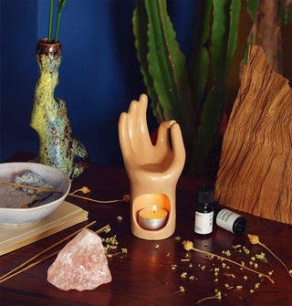Mudra hand oil diffuser with lid tea light in its base sits on a wooden tabletop with dried flowers, a large crystal, essential oil bottles, and other artful items