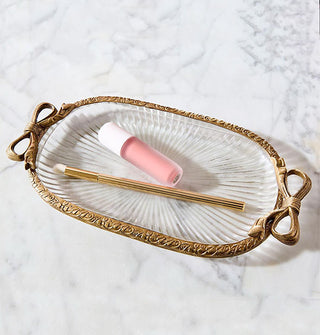 Oval-shaped ribbed glass dish with brass rim featuring bow accents on either side holds a makeup brush and lip gloss tube
