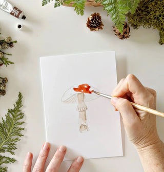 Model paints a mushroom on a white card surrounded by moss, ferns, and pinecones