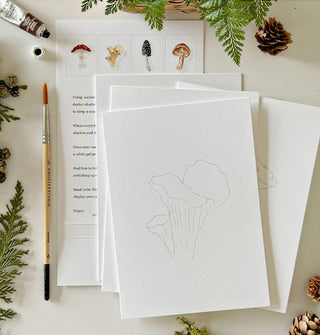 White notecards with mushroom outlines to be painted in with instruction card underneath, all flanked by ferns, pinecones, paintbrush, and tube of paint