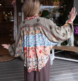 Model facing away from the camera with arms upraised wears a loose-fitting shirt with patchworked floral prints, billowy sleeves, and floral embroidery across the back and arms