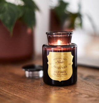 A lit Apothecary Noir Paddywax Palo Santo candle shines from inside a black apothecary-style jar on a wooden surface with houseplants in the background