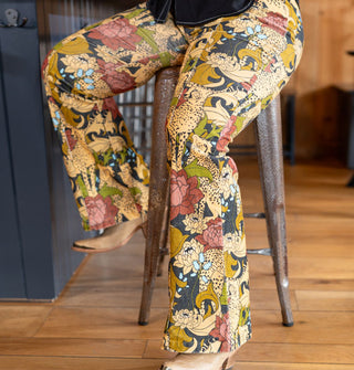 Model sitting on a bar stool wears a pair of pants in a cheetah and floral print with a black button-down shirt and tan cowgirl boots