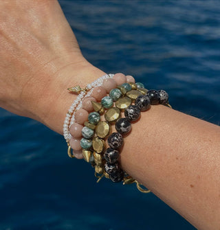 Model wears an assortment of brass and gemstone beaded bracelets against a backdrop of blue water