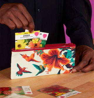 Model pulls flower seed packets from a rectangular pouch with red top zipper that features a colorful illustration of hummingbirds gathered around a hibiscus flower