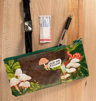 Wristwatch, bandage, and ballpoint pen spill out of a rectangular pouch with green top zipper that features an all-over design of Bigfoot's feet in a grassy forest floor with mushrooms and a gnome who's saying, "You're kind of a legend"