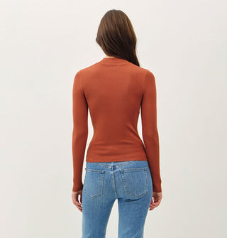Model facing away from the camera wears a long-sleeved rust-colored mock turtleneck top with blue jeans