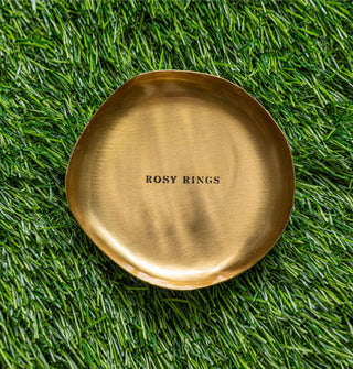 Small rounded gold-toned dish stamped with the words, "Rosy Rings" is placed on green astroturf