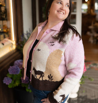 Smiling model wears a button-down collared graphic cardigan with a pink, white, tan, and brown desert landscape scene