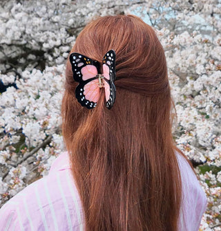 Model standing in front of a backdrop of white blossoms wears a pink monarch butterfly clip in a partially swept-back hairstyle
