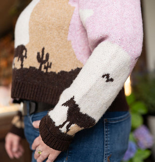 Model wears a graphic sweater with a pink, white, tan, and brown desert landscape scene