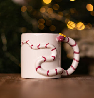 White coffee mug features a three-dimensional pink striped snake with a yellow flower on its head slithering across the surface and forming a handle at one side