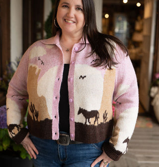 Smiling model wears a button-down collared graphic cardigan with a pink, white, tan, and brown desert landscape scene